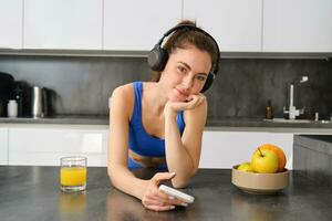 retrato do jovem ginástica mulher com fones de ouvido, bebendo laranja suco dentro cozinha e usando Smartphone, ouvindo música, obtendo pronto para exercite-se Academia foto