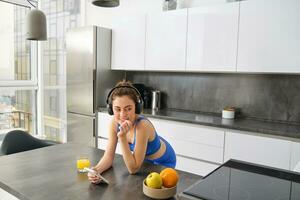 estilo de vida e dar certo. jovem sorridente mulher dentro fones de ouvido, em pé dentro cozinha com Smartphone, bebendo laranja suco e ouvindo música, posição sobre para academia, indo corrida foto