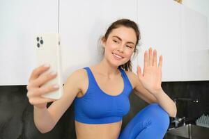 retrato do jovem ginástica garota, social meios de comunicação blogueiro, viver transmissão, dizendo Olá às Smartphone, acenando mão e sorridente, vestindo roupa ativa foto