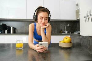 retrato do jovem ginástica mulher com fones de ouvido, bebendo laranja suco dentro cozinha e usando Smartphone, ouvindo música, obtendo pronto para exercite-se Academia foto
