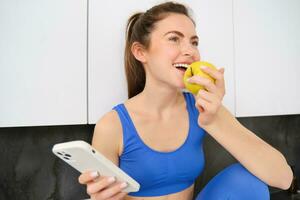 imagem do à moda desportista, jovem ginástica instrutor sentado dentro cozinha e comendo a maçã, segurando Smartphone, usando social meios de comunicação aplicativo em Móvel telefone foto