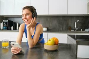 imagem do feliz, à moda jovem Esportes mulher, em pé dentro cozinha e bebendo laranja suco, ouvindo música dentro fones de ouvido, usando Smartphone aplicativo foto