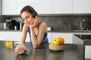 retrato do jovem ginástica mulher com fones de ouvido, bebendo laranja suco dentro cozinha e usando Smartphone, ouvindo música, obtendo pronto para exercite-se Academia foto