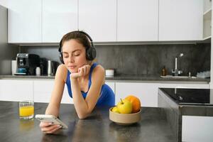 imagem do feliz, à moda jovem Esportes mulher, em pé dentro cozinha e bebendo laranja suco, ouvindo música dentro fones de ouvido, usando Smartphone aplicativo foto