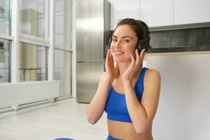 imagem do ginástica menina fazendo Esportes às lar, coloca em sem fio fones de ouvido para música durante exercite-se Treinamento sessão, vestindo azul sportsbra foto