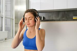 jovem mulher coloca em sem fio fones de ouvido para ouço música durante exercite-se às lar, desgasta sutiã esportivo, sorrisos com satisfeito face foto