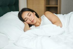 lindo ásia menina dorme dentro dela cama com branco folhas, caloroso inverno edredon, sorridente dentro dela dormir, sonhando, deitado dentro quarto dentro manhã foto