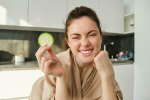 retrato do lindo morena menina cozinhando dentro a cozinha, posando dentro roupão de banho às lar, segurando abobrinha, mostrando feliz sorriso, fazer saudável comida, vegetariano refeição foto