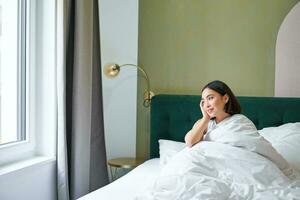 feliz coreano mulher acorda acima dentro dela hotel sala, deitado dentro cama debaixo acolhedor caloroso cobertores, branco folhas, desfrutando final de semana manhã foto