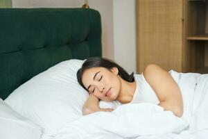 lindo ásia menina dorme dentro dela cama com branco folhas, caloroso inverno edredon, sorridente dentro dela dormir, sonhando, deitado dentro quarto dentro manhã foto