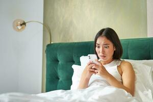 retrato do chocado coreano mulher olhando às dela Móvel telefone, deitado dentro cama, lendo mau chocante notícia em Smartphone aplicativo foto