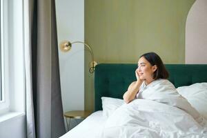 lindo sonolento ásia menina acorda acima dentro dela cama com acolhedor branco cobertores, olhando lado de fora janela com satisfeito sorriso. estilo de vida conceito foto