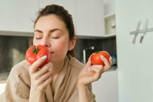 retrato do lindo jovem mulher cheirando tomates, cozinhando salada a partir de fresco legumes comprou dentro mercado, preparando Comida para família, conduzir ativo e saudável estilo de vida, em pé dentro cozinha foto