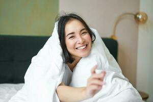 lindo ásia mulher sentado em cama, coberto com branco edredon, sorridente, desfrutando feliz final de semana manhã, rindo às Câmera foto