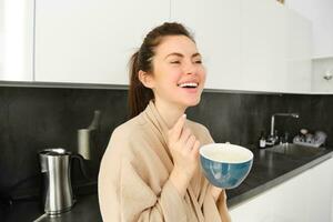 retrato do boa aparência jovem mulher iniciando dela dia com copo do café, em pé dentro a cozinha e bebendo cappuccino a partir de grande caneca, desfrutando favorito beber dentro a manhã foto