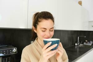 retrato do lindo jovem mulher dentro roupão de banho, bebendo manhã café e desfrutando a gosto, sorridente satisfeito, em pé dentro a cozinha foto