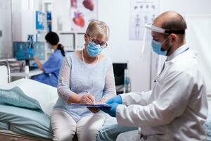 Senior mulher assinatura médico teste resultados durante consulta com médico dentro hospital exame quarto e vestindo face mascarar contra cobiçado para segurança precaução. foto