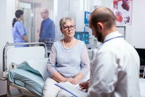 Senior mulher respondendo médico questões durante exame dentro hospital sala. converstation com médico coisa clínica remédio assistência médica, Senior, casaco, prancheta. foto