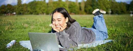 sorridente ásia menina deitado dentro parque em grama, assistindo filme ou vídeo em computador portátil, olhando às tela com interesse foto