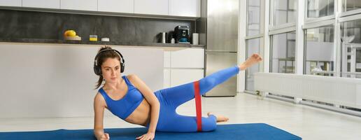 imagem do desportivo menina fazendo aeróbica, usando resistência banda em pernas, exercite-se às lar, escuta para música dentro fones de ouvido foto