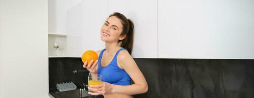 interior tiro do mulher depois de dar certo, em pé dentro cozinha com fresco suco e a laranja, bebendo isto foto