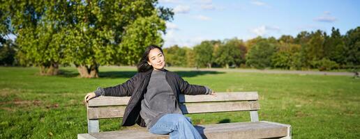 retrato do jovem mulher dentro ao ar livre roupas, sentado em Banco relaxado, sorridente e desfrutando Visão em verde parque foto