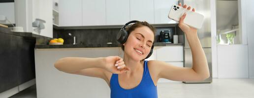 felicidade, Esportes e bem-estar. jovem mulher dançando dentro fones de ouvido, segurando Smartphone, fazendo exercite-se às lar, ginástica Treinamento dentro dela casa foto