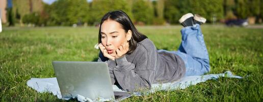 retrato do lindo menina deitado dentro parque e olhando com surpreso face às dela computador portátil, assistindo vídeos, conversando com amigos enquanto relaxante ao ar livre foto