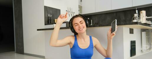 alegre morena menina faz ginástica às lar, desgasta sem fio fones de ouvido e danças, detém Smartphone, faz exercite-se dentro de casa em chão, desgasta azul roupa ativa foto