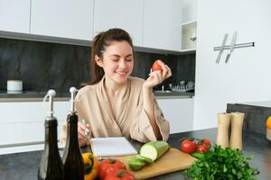 retrato do lindo jovem mulher dentro a cozinha, escrevendo baixa cozinhando receita, sentado perto cortar borda com legumes e fazer mercearia lista, criando saudável vegetariano cardápio para dela família foto