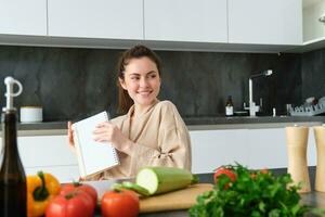 retrato do mulher escrevendo baixa Lista do mantimentos, fazer notas dentro receita, sentado dentro cozinha perto vegetais, preparando jantar cardápio foto