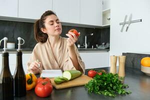 retrato do lindo sorridente mulher, escrevendo dela saudável cardápio, comendo tomate enquanto culinária, fazer mercearia lista, sentado dentro a cozinha foto