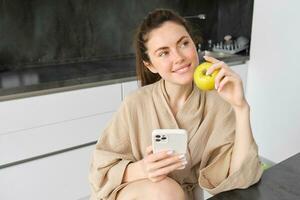 fechar acima retrato do feliz jovem mulher dentro roupão de banho, sentado dentro a cozinha e usando Móvel telefone, segurando a maçã, ordem frutas e legumes on-line, usando Smartphone aplicativo para mercearias Entrega foto