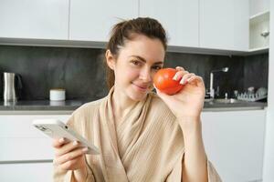 fechar acima retrato do lindo sorridente mulher, segurando fresco tomate, sentado dentro cozinha com Smartphone, ordens legumes on-line, usando inscrição para Comprar mantimentos, usando Móvel telefone foto