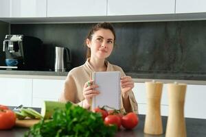 retrato do feliz jovem mulher escreve baixa cardápio para jantar, senta dentro a cozinha perto vegetais, faz mercearia Lista para compras, poses dentro a roupão de banho foto