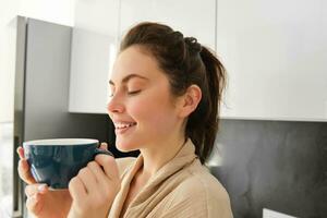retrato do feliz jovem mulher começa dela manhã com caneca do café, bebendo chá a partir de xícara, em pé dentro a cozinha, sorridente alegremente foto