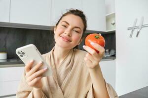 fechar acima retrato do lindo sorridente mulher, segurando fresco tomate, sentado dentro cozinha com Smartphone, ordens legumes on-line, usando inscrição para Comprar mantimentos, usando Móvel telefone foto