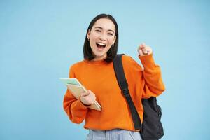 alegre ásia estudante, menina com mochila e cadernos, indo para faculdade, parece feliz, azul fundo foto