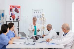 médico Senior mulher segurando osso estrutura discutindo humano estrutura usando corpo esqueleto apresentando cuidados de saúde tratamento. hospital grupo trabalhando às médico perícia dentro encontro quarto foto