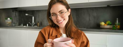 fechar acima retrato do lindo menina dentro copos, segurando quente copo do bebida, bebendo chá ou café às casa e sorridente às Câmera foto