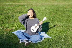 animado ásia menina senta dentro parque com cavaquinho, tocam instrumento e sente feliz otimista, positivo momentos e pessoas conceito foto
