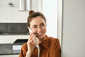 retrato do feliz mulher às lar, respostas telefone chamar, falando em móvel, segurando Smartphone e sorridente foto