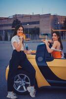 bonita fêmeas dentro casual roupas comendo pizza, posando dentro amarelo carro com francês fritas e refrigerante dentro vidro garrafa em Está porta-malas. velozes Comida. cheio comprimento foto