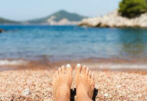 pés do jovem mulher com pedicure em uma seixo de praia em período de férias. desfrutando a Sol em uma verão ensolarado dia às a mar. período de férias e viagem conceito. fechar-se. foto