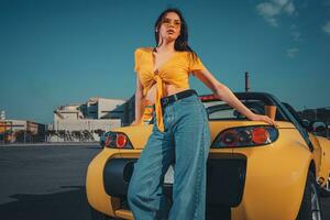 fêmea dentro azul jeans, laranja topo e oculos de sol é posando perto amarelo carro cavalo estradeiro com papel copo do chá em tronco às estacionamento muito. cópia de espaço foto