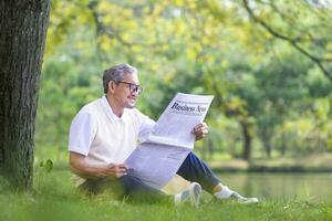 Senior ásia homem lendo uma o negócio jornal enquanto sentado debaixo a árvore de a lago às a público parque para lazer, lazer e relaxamento dentro natureza foto