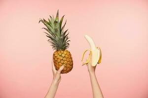 cortada foto do jovem mulher elevado mãos com nu manicure segurando fresco abacaxi e descascado banana enquanto ser isolado sobre Rosa fundo