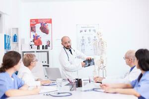 cirurgião segurando radiografia e fazer apresentação dentro frente do colegas dentro hospital encontro sala. clínica especialista terapeuta falando com colegas de trabalho sobre doença, remédio profissional foto