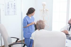 fêmea enfermeira fazendo apresentação do humano anatomia em esqueleto dentro frente do médicos. clínica especialista terapeuta falando com colegas sobre doença, remédio profissional foto
