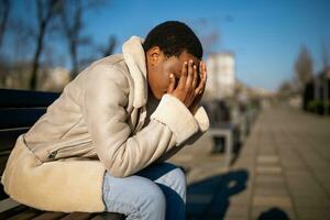 ao ar livre retrato do depressivo afro-americano mulher em ensolarado dia. ela é sentado em Banco dentro a rua. foto